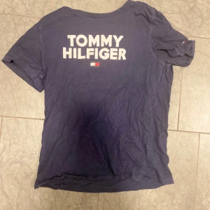Marinblå Tommy Hilfiger t-shirt - Snygg marinblå t-shirt från Tommy Hilfiger i ekologisk bomull. Stor logga och flagga tryckt på ryggen, klassisk passform och korta ärmar. Perfekt för dig som gillar stilrena och ikoniska plagg med streetkänsla.