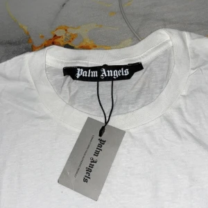 Vit Palm Angels t-shirt London print - Snygg vit t-shirt från Palm Angels med klassiskt London-tryck framtill i svart och lila. T-shirten har rund hals, korta ärmar och är gjord i mjuk bomull. Perfekt för dig som gillar streetwear och vill sticka ut med en ikonisk logga.