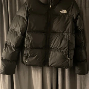 Svart pufferjacka från The North Face - Säljer en svart pufferjacka från The North Face med klassisk logga på bröstet. Jackan har en boxig passform, dragkedja framtill och två sidofickor. Perfekt för kalla dagar och riktigt snygg till streetwear-stilen.