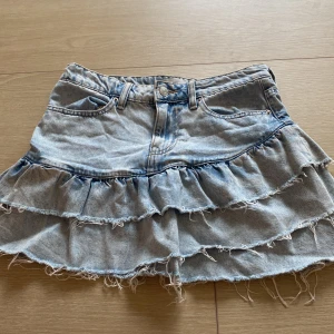 Volang Jeanskjol  - Säljer en ljusblå jeanskjol från Gina tricot i storlek 34. Kjolen har volanger, fransiga kanter och klassiska jeansdetaljer som fickor och bälteshällor. Är low waist.