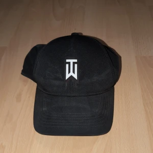Nike Tiger Woods svart keps Dri-FIT - Svart keps från Nike Tiger Woods Collection med vit TW-logga framtill och vit Nike Swoosh bak. Modellen är Legacy91 med Dri-FIT ADV-teknologi och perforerad baksida för extra ventilation. Perfekt för sport och träning, stilren och funktionell.