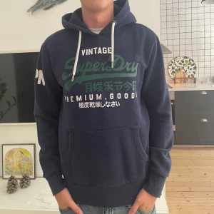 Blå Superdry hoodie med tryck - Mörkblå hoodie från Superdry med grönt och vitt tryck framtill och texten JPN på ärmen. Klassisk känguruficka, vita snören i huvan och ribbade muddar. Perfekt för dig som gillar streetwear och vill ha en snygg och bekväm tröja.