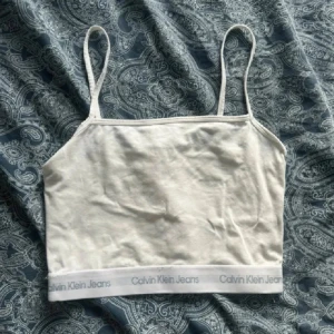 Vit linnetopp från Calvin Klein Jeans - Säljer en vit linnetopp från Calvin Klein Jeans med smala axelband och elastisk kant med logotyp. Toppen är croppad och har en enkel, stilren design som passar perfekt till jeans eller shorts. Materialet är mjukt och bekvämt bomull.