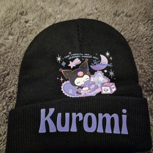 Svart Kuromi mössa med tryck - Svart stickad mössa med stort lila Kuromi-tryck framtill och söt illustration av Kuromi bland stjärnor och måne. Mysig och trendig accessoar för dig som gillar kawaii-stil och Sanrio-karaktärer.