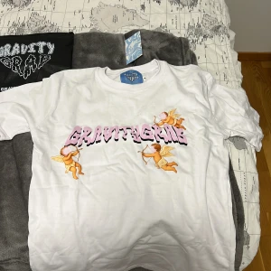 Vit Gravity Grab t-shirt med änglar - Vit t-shirt från Gravity Grab med tryck i rosa och svart text samt gulliga änglar på framsidan. Klassisk passform och rund halsringning. Perfekt för dig som gillar streetwear och vill sticka ut med ett unikt motiv.
