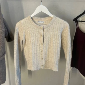 Beige kabelstickad kofta Holly & Whyte - Superfin beige kabelstickad kofta från Holly & Whyte by Lindex. Croppad modell med rund halsringning och snygga knappar framtill. Perfekt till lager-på-lager och ger en mysig vibe till din outfit.