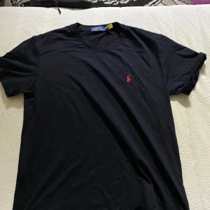 Svart t-shirt från Polo Ralph Lauren - Storlek XXL men passar som XL/L. Svart t-shirt från Polo Ralph Lauren med klassisk passform och rund hals. Ikoniska röda Polo-loggan broderad på bröstet. Tillverkad i mjuk bomull som känns skön mot huden. Perfekt för en clean och stilren look.