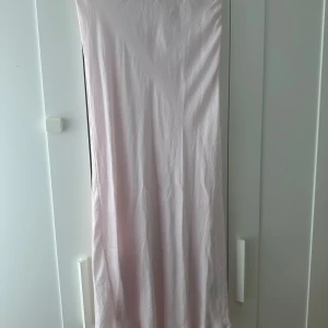 Ljusrosa långkjol i satin - En stilren och enkel ljusrosa långkjol i mjukt satinliknande material. Klänningen har raka linjer och faller snyggt längs kroppen. Perfekt för dig som gillar minimalistisk och elegant stil.