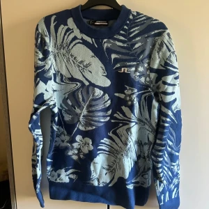 J linderberg tröja  - Snygg blå stickad tröja från J.Lindeberg med ljusblått blommigt mönster och logga på bröstet. Rund hals och långa ärmar. Perfekt för dig som gillar att sticka ut med coola prints och vill ha något som känns både trendigt och bekvämt.