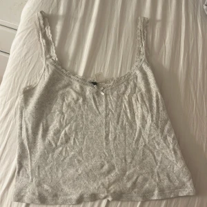 Vit spetsdetaljerad linnetopp H&M Divided - Supersöt vit linnetopp från H&M Divided med spetskant runt axelband och halsringning. Toppen har en liten rosett framtill och är ribbad med diskret hålmönster. Perfekt för lager-på-lager eller sommaroutfits.