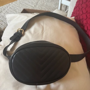 Svart midjeväska med guld-detaljer - Snygg svart midjeväska i skinnimitation med justerbart bälte på 13 lägen och guldigt spänne. Väskan har oval form, dragkedja i guld och diskret mönster i v-format. Perfekt för att bära det viktigaste och ge en trendig touch till din outfit.