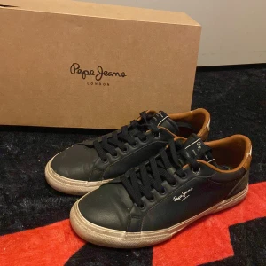 Blåa sneakers från Pepe Jeans - Blåa  sneakers från Pepe Jeans med vita sulor och bruna detaljer runt hälen. Skorna har snörning, rund tå och logga på sidan samt plösen. Materialet är en mix av skinn och syntet. Snygg kontrast mellan blåa, vitt och brunt för en clean streetstil.