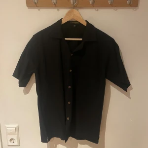 Svart kortärmad skjorta Ton Mon Clothing - Svart skjorta från Ton Mon Clothing i storlek M. Skjortan har korta ärmar, klassisk krage och knäppning med bruna knappar framtill. Enkel och stilren design som passar perfekt till jeans eller shorts. Materialet känns lätt och bekvämt mot huden.