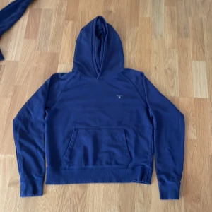Blå hoodie från Gant med logga - Säljer en stilren marinblå hoodie från Gant med liten broderad logga på bröstet. Tröjan har huva, magficka och långa ärmar. Perfekt för dig som gillar klassisk och enkel streetstil.