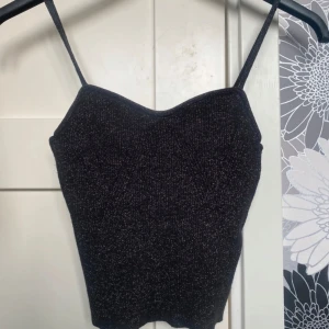 Glittrig svart linne från H&M - Säljer en svart glittrig linne från H&M i storlek S. Toppen har smala axelband och en snygg croppad passform med lätt v-ringad hals. Materialet är mjukt och stretchigt med glittriga trådar som ger en cool effekt. Perfekt för att lyfta din outfit!