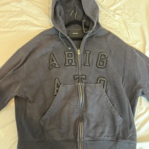 Grå hoodie från Axel Arigato - Säljer en grå hoodie från Axel Arigato med broderad text framtill. Tröjan har dragkedja, huva med snörning och en stor magficka. Perfekt för dig som gillar streetwear och oversized look.