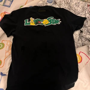 Svart Lacoste t-shirt med krokodillogo - Svart t-shirt från Lacoste i bomull med klassisk rund hals och coolt tryck av krokodil och Lacoste-text på bröstet samt ett större grafiskt tryck på ryggen. Regular fit och korta ärmar. Perfekt för dig som gillar streetwear och snygga loggor.