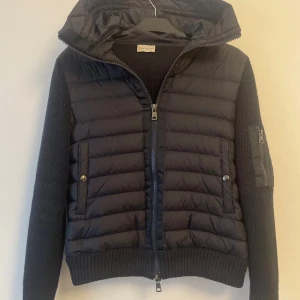 Svart Moncler pufferjacka med stickade ärmar/Moncler Cardigan  - Svart pufferjacka från Moncler med stickade ärmar och huva. Jackan har quiltad front, ribbade muddar och tvåvägsdragkedja. Ärmarna är prydda med Moncler-logga och en ficka med dragkedja. Perfekt för dig som vill ha en snygg och sportig look.