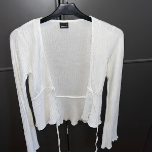 Vit ribbad kofta från Gina Tricot - Säljer en vit ribbad kofta från Gina Tricot i storlek S. Koftan har långa ärmar, knytband framtill och vågig nederkant. Perfekt att slänga över ett linne eller topp för en chill och trendig look.