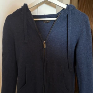 Mörkblå stickad hoodie - Mysig mörkblå stickad hoodie från Gutteridge i kashmirblandning. Tröjan har dragkedja framtill, två fickor och huva med snörning. Liknar soft goat tröjorna. Finns inga nopprar och är som nyskick. Funkar för både män och kvinnor. Skriv för mer info
