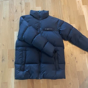 Svart pufferjacka från Sail Racing - Säljer en svart pufferjacka från Sail Racing med hög krage och dragkedja framtill. Jackan har två stora fickor, diskret logga på bröstet och en rund logga på ryggen. Perfekt för kalla dagar och riktigt skön att bära. Storlek 176 men är stor så sitter som en S