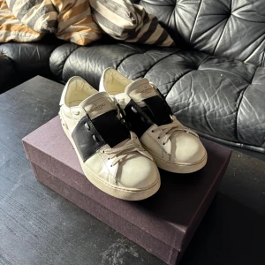 Valentino Garavani Open sneakers vit/svart - Säljer ett par Valentino Garavani Open sneakers i vit skinn med bred svart läderdetalj över snörningen. Klassisk rund tå och platt sula, med vita snören och subtila logodetaljer på plösen. Snygg och stilren design som sticker ut. Priset kan diskuteras vid snabb och enkel affär.