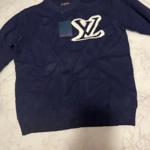 Marinblå stickad tröja Louis Vuitton - Snygg marinblå stickad tröja från Louis Vuitton med stort vitt LV-logo på bröstet. Tröjan har rund halsringning och långa ärmar, perfekt för dig som vill ha en exklusiv och trendig look.