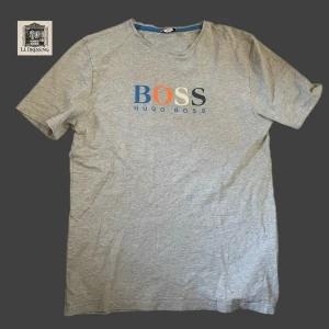 Grå t-shirt från Hugo Boss, storlek M - Snygg grå t-shirt från Hugo Boss i storlek M. T-shirten har korta ärmar och rund hals. Framsidan pryds av BOSS-loggan i blått, orange, vitt och mörkblått. Tillverkad i mjuk bomull som är skön mot huden. Perfekt för en chill och stilren look.