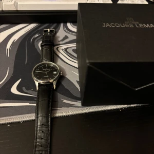 Jacques Lemans svart herrklocka - Snygg automatisk klocka från Jacques Lemans med svart urtavla och datumvisning. Klockan har ett klassiskt runt silverfärgat boett och ett svart läderarmband med krokodilpräglad yta. Perfekt för dig som gillar stilrena accessoarer.