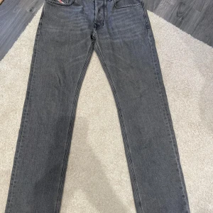 Grå jeans från Diesel - Säljer ett par grå jeans från Diesel med klassisk femficksdesign och raka ben. Jeansen har snygga detaljer vid fickorna och en diskret tvättad look. Materialet är jeans i bomull och passformen är normal. Perfekta för dig som gillar stilrena och tidlösa jeans.