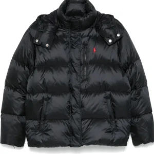 Svart pufferjacka från Polo Ralph Lauren - Säljer en svart pufferjacka från Polo Ralph Lauren med röd logga på bröstet. Jackan har hög krage, dragkedja framtill och praktiska fickor. Perfekt för kalla dagar och riktigt snygg till streetwear-stilen.