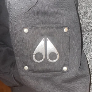 Svart vinterjacka Moose Knuckles S - Svart vinterjacka från Moose Knuckles med vit teddyfoder i huvan. Jackan har quiltat innerfoder med logomönster, metallogo på ärmen och praktiska fickor. Perfekt för kalla dagar och riktigt snygg streetstil.