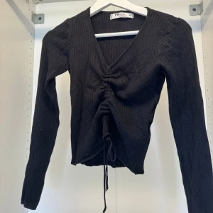Svart ribbad topp med snörning - Svart långärmad topp från New Yorker i ribbad stickad polyester. V-ringad med snygg snörning framtill som ger en croppad look. Perfekt för dig som gillar stilrena och trendiga plagg med lite edge.