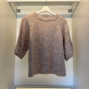 Beige stickad tröja med halvärm - Mysig beige stickad tröja med halvärm och rund hals. Tröjan har en avslappnad passform och ribbade detaljer vid hals, ärmslut och nederkant. Perfekt för lager-på-lager under hösten eller våren.