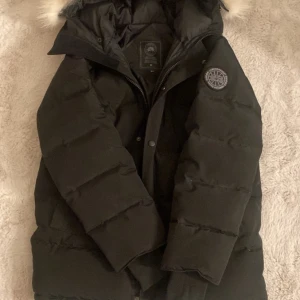 Svart dunjacka från Canada Goose - Svart Canada Goose vinterjacka med äkta vargpäls på huvan. Jackan är i mycket fint skick, varm och bekväm med klassisk design. Passar både vardag och kallare väder. Originalmärke och etikett finns kvar.