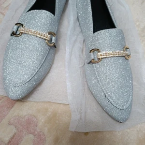 Glittriga silverloafers med pärldetalj - Snygga loafers i glittrigt silver med spetsig tå och platt sula. Dekorerade med guldfärgade spännen och pärlor framtill för extra bling. Perfekta för dig som vill sticka ut med stil och gillar glam. Skorna har en elegant form och passar till många olika looks.