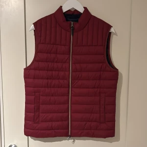 Röd dunväst från Hackett London - Snygg röd dunväst från Hackett London med ståkrage och horisontella quiltade sömmar. Västen har två fickor framtill och dragkedja hela vägen. Materialet är lätt och funktionellt, tillverkat av 100% återvunnet syntet. Perfekt för lager-på-lager under kyliga dagar.