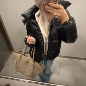 Svart pufferjacka med dragkedja - Säljer en svart, glansig pufferjacka med hög krage och dragkedja framtill. Jackan har en quiltad design och är perfekt för kyligare dagar. Passformen är normal och jackan har långa ärmar. Enkel och stilren modell som funkar till många outfits.