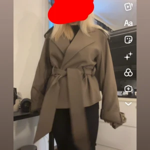 Brun/grå trenchcoat med bälte - Snygg brun/grå trenchcoat med breda slag och knytskärp i midjan. Jackan har långa ärmar med spänndetaljer vid handlederna och en loose passform. Använd 1 gång så i super bra skick