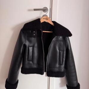 Svart teddy-skinnjacka med dragkedja - Säljer en svart jacka med teddyfoder och detaljer i skinn. Jackan har bred krage, muddar och nederkant i teddy, samt praktisk dragkedja framtill. Snygg och trendig modell som passar perfekt till kalla dagar.