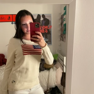 Vit stickad tröja från Ralph Lauren - Vit stickad tröja från Ralph Lauren med amerikansk flagga broderad på bröstet och RL-broderi vid nederkanten. Tröjan har rund halsringning och långa ärmar, perfekt för en clean och klassisk look.