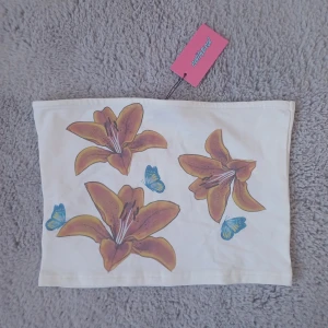 Vit blommig tubtopp från Ediktead - Supersöt vit tubtopp från Edikted med stora orange-gula liljor och blå fjärilar på framsidan. Bandeau-modell som sitter snyggt över bröstet. Perfekt för dig som vill ha en unik och färgglad topp till sommaren. Materialet känns mjukt och stretchigt.