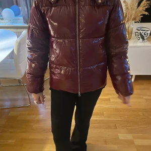 Vinröd pufferjacka från Nelly XS - Snygg vinröd pufferjacka från Nelly i glansigt material. Jackan har huva, dragkedja framtill och quiltad design. Perfekt för kalla dagar och ger en cool vibe till din outfit.