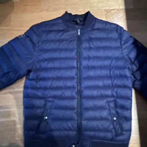 Mörkblå pufferjacka Polo Ralph Lauren XL - Snygg mörkblå pufferjacka från Polo Ralph Lauren i storlek XL. Jackan har quiltad design, dragkedja framtill, två fickor med tryckknapp och en patch med Polo-logga på ärmen. Perfekt för kyliga dagar och riktigt clean stil.