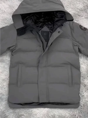 Canada Goose  - Snygg grå pufferjacka med huva och diskret logga på ärmen. Jackan har dragkedja och tryckknappar framtill samt två sidofickor. Perfekt för kalla dagar och ger en clean look.