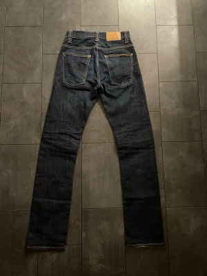 Mörkblå jeans från Nudie Jeans Co - Säljer ett par mörkblå jeans från Nudie Jeans Co med klassisk femficksdesign och kontrastsömmar i orange. Jeansen har rak passform och är tillverkade i slitstarkt denimtyg av bomull. Märkespatch i läder baktill och snygga detaljer på bakfickorna.kan gå ner i pris vid snabb affär 