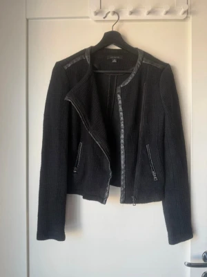 Svart cardigan jacka med skinn-detaljer - Svart cardigan jacka från Ann Taylor med coola skinn-detaljer längs axlar och kanter. Jackan har långärmad design, dragkedjor på fickorna och en croppad passform som ger en modern vibe. Perfekt för dig som gillar stilrena och edgy plagg.