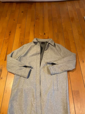 Grå kappa från Vero Moda, storlek M - Säljer en stilren grå kappa från Vero Moda i storlek M. Jackan har klassisk krage, raka linjer och tryckknappar framtill. Materialet är mjukt och känns som ullblandning. Perfekt för dig som gillar minimalistisk och clean look. Lång rock.