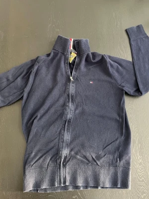 Marinblå kofta från Tommy Hilfiger - Säljer en marinblå kofta från Tommy Hilfiger med dragkedja framtill och ståkrage med röd och vit detalj. Liten logga broderad på bröstet. Tröjan är långärmad och har ribbade muddar vid ärmslut och nederkant. Perfekt för kyliga dagar. Hör av er vid frågor!
