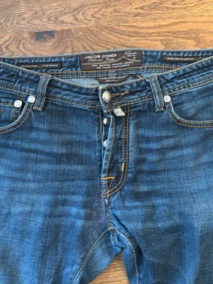 Jacob Cohen blå jeans med raka ben - Snygga blå jeans från Jacob Cohen med klassisk femficksdesign och raka ben. Jeansen har kontrastsömmar, knappgylf och är tillverkade i mjukt bomullsmaterial. Perfekt för dig som gillar stilrena och tidlösa jeans med exklusiv känsla.